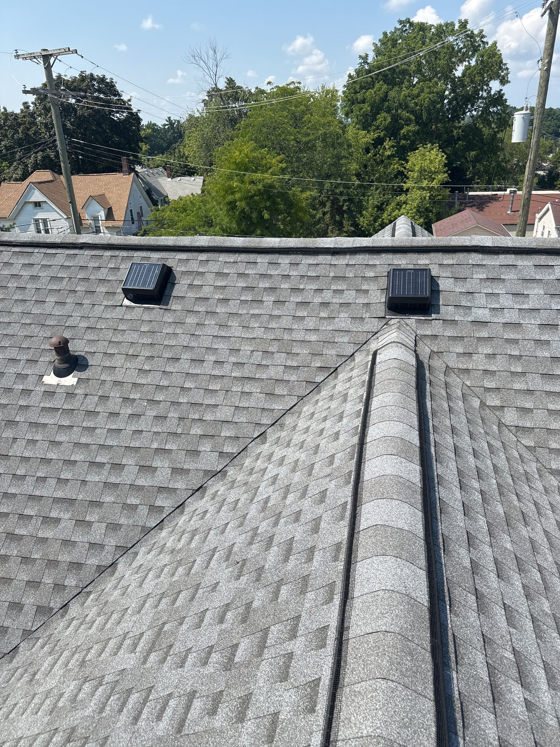 Gray asphalt shingle roof with two black vents, ridge cap, and a small metal pipe. Blue sky background.