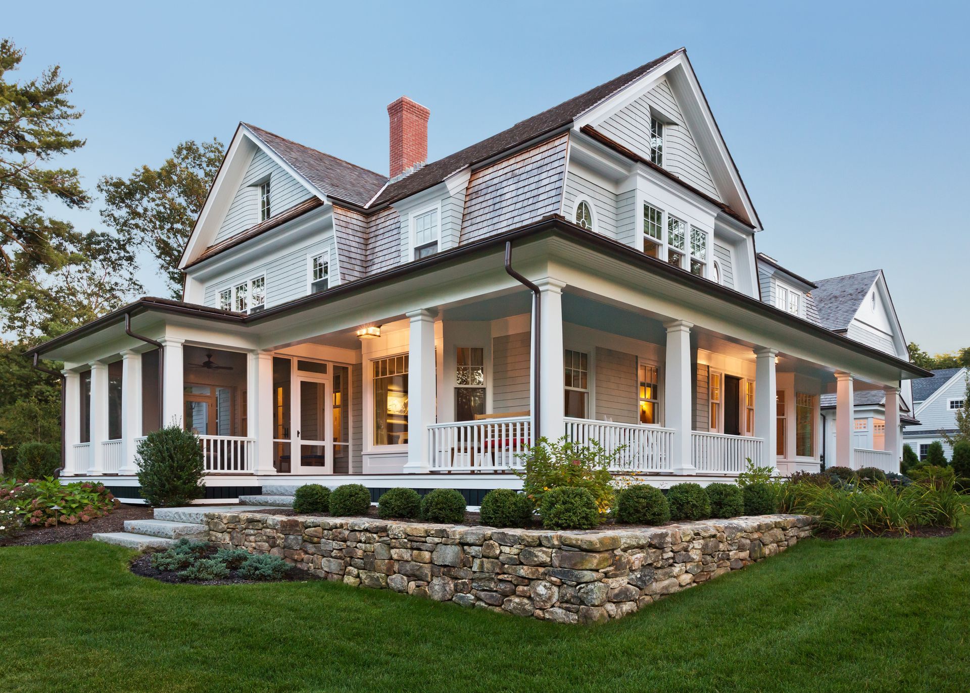 Two-story light gray house with wraparound porch and stone retaining wall; lush green lawn.