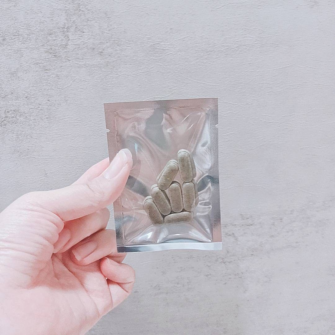 一手拿著一個密封的銀色小袋，裡面裝著幾粒長方形的淺棕色營養補充膠囊。