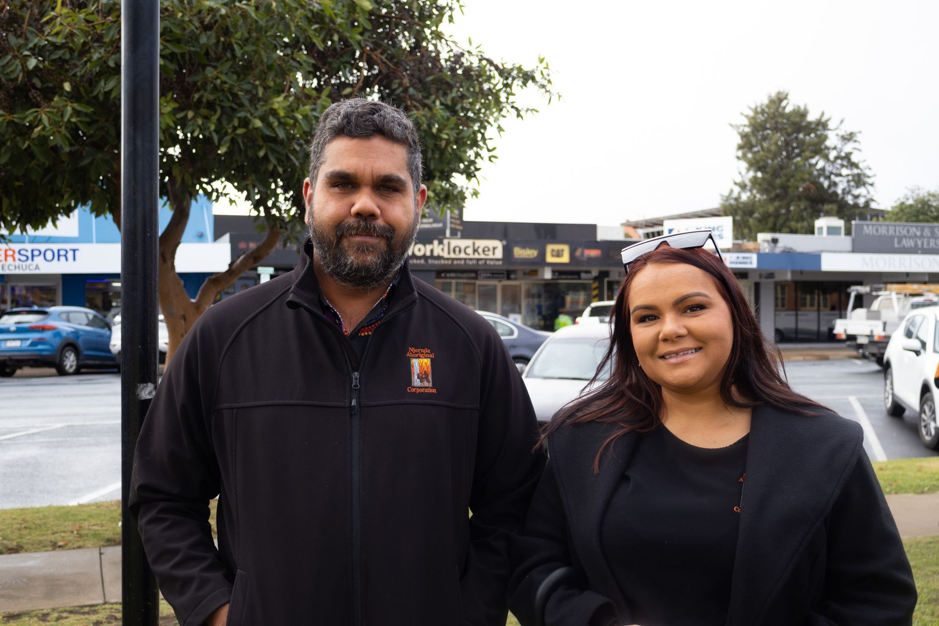 A man and woman stand side-by-side outdoors. The man wears a black jacket and looks at the camera. The woman smiles. Behind them are shops and cars.