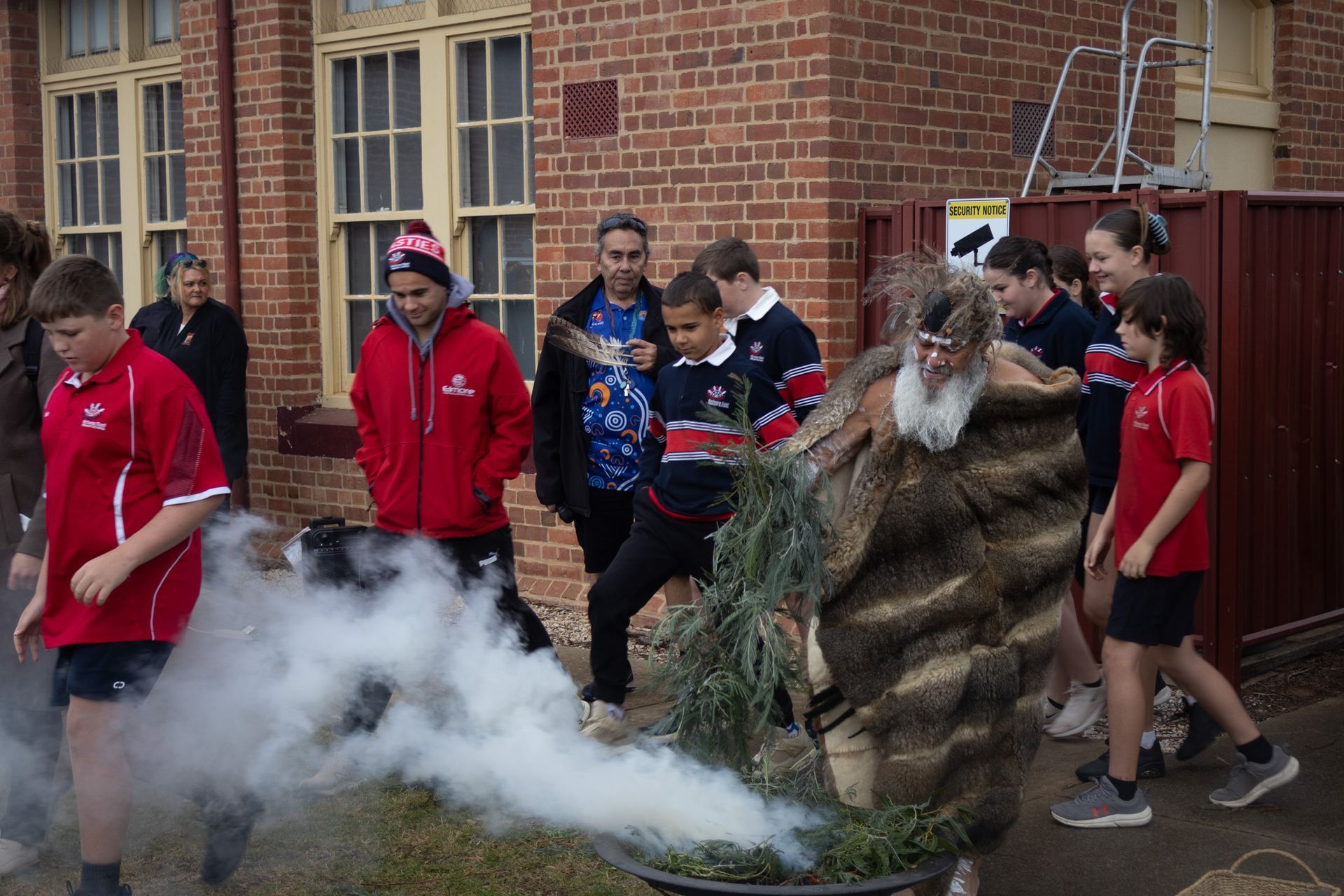 Children and adults watch as a person in traditional attire smolders native foliage creating smoke.  Group stands outdoors near a brick building.