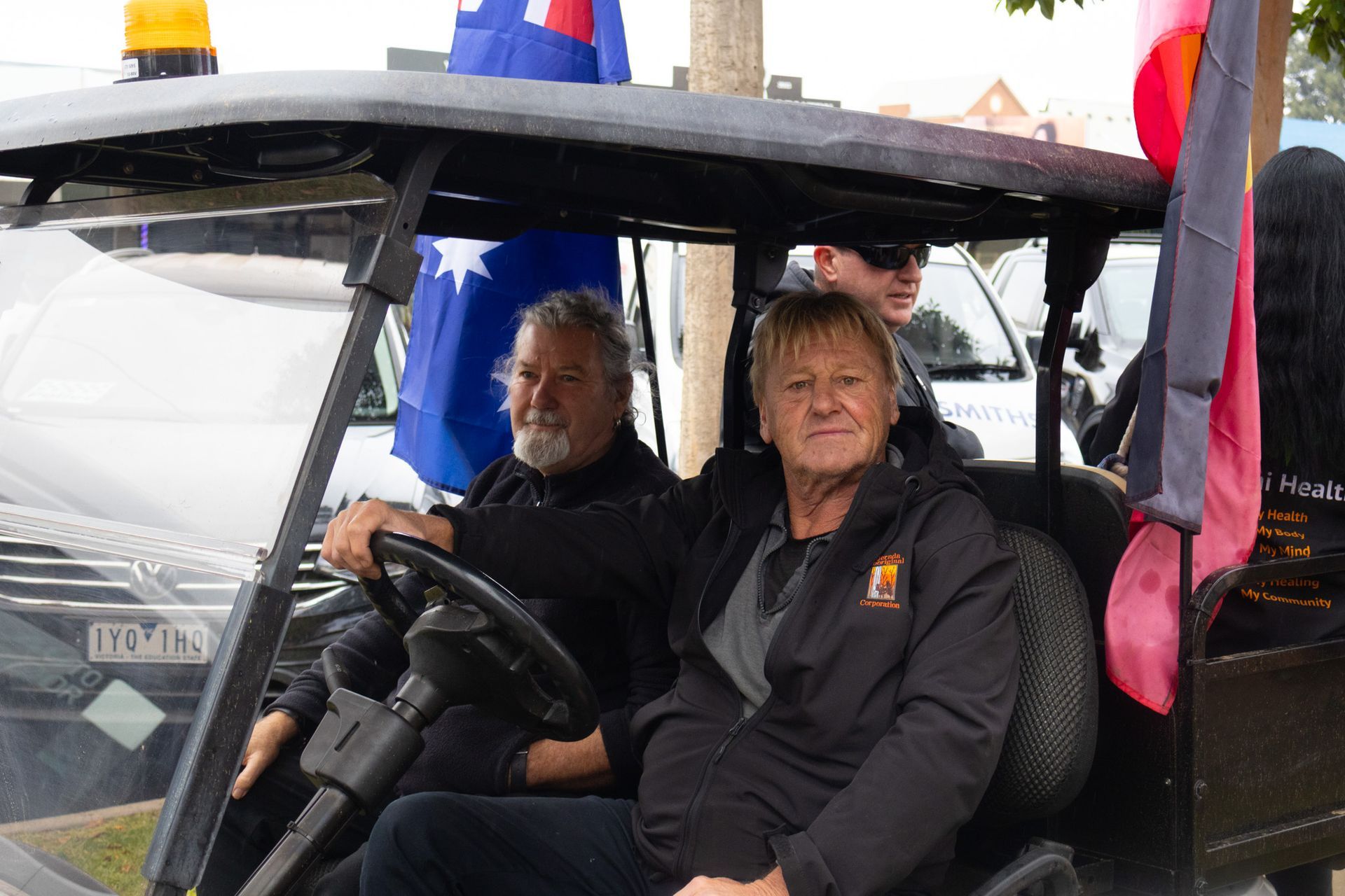 Two people in a golf cart, Australian flags visible. One drives, the other smiles; a third person sits in the back.