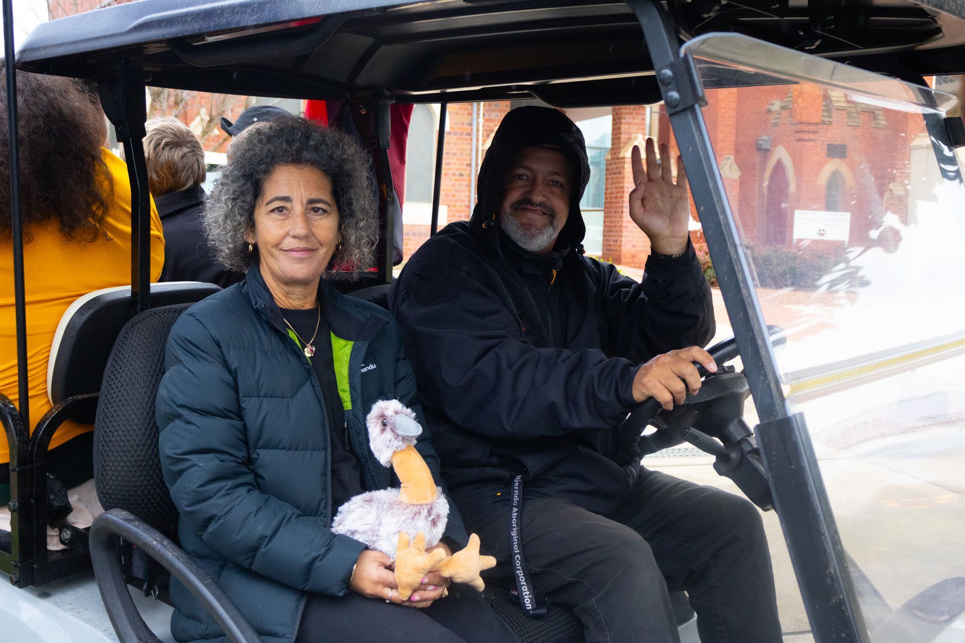 Two people in a golf cart; a woman holding a stuffed bird smiles, while a man waves. They are outside with a building in the background.