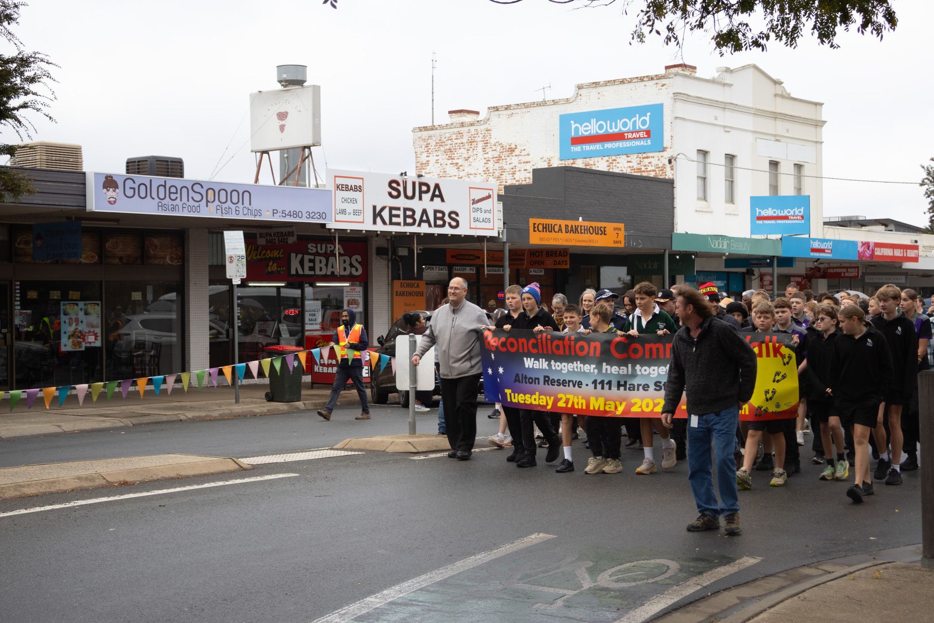 A group of people march down a street, holding a banner. Shops line the road under an overcast sky.