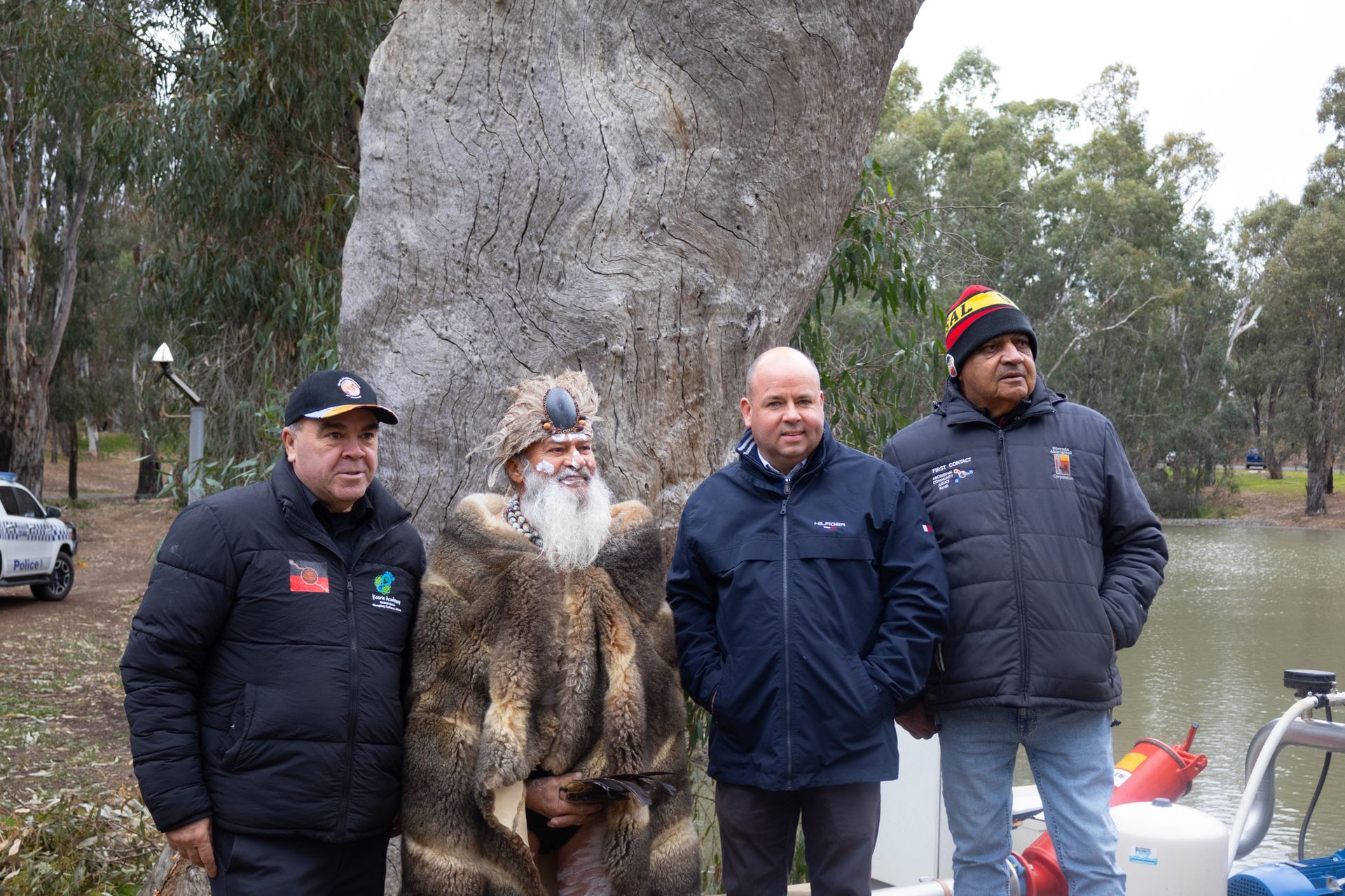 Four men stand outdoors by a river, one in fur, others in jackets. Trees and a cloudy sky are in the background.