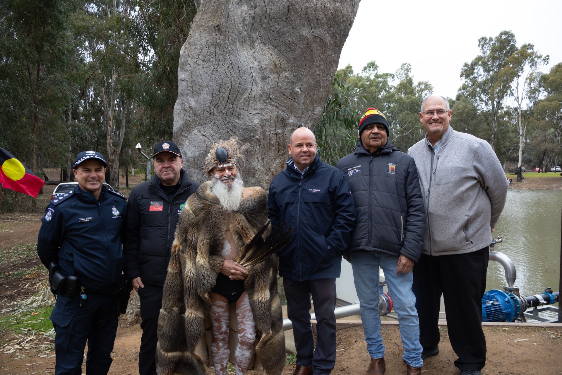 A group of six people posing outdoors near a large tree and a body of water. The group includes a police officer and a person wearing a traditional fur garment.