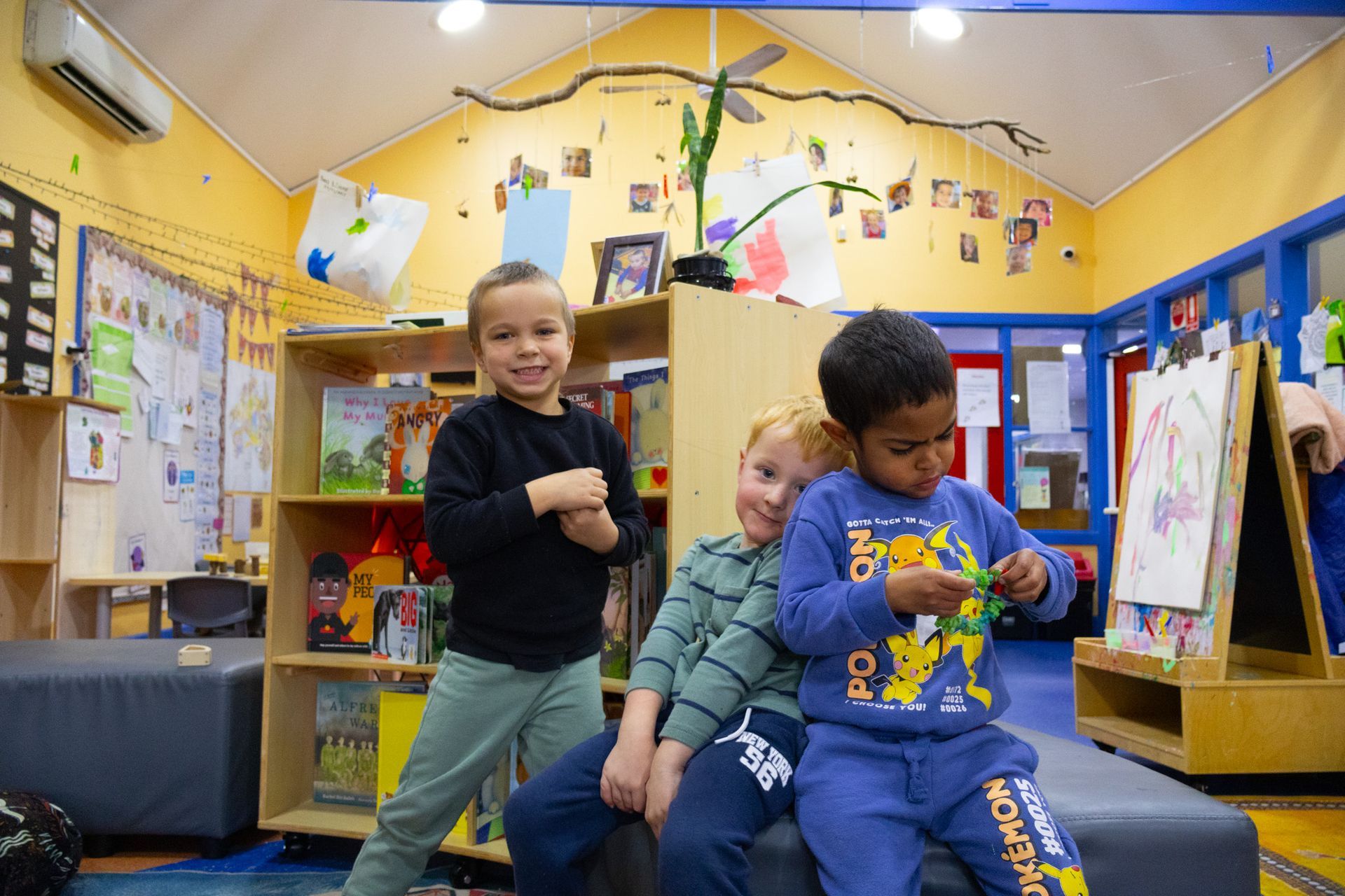 Three young children smiling and interacting in a colorful classroom setting. One boy stands, while the other two sit near a bookshelf, and one boy holds something green.