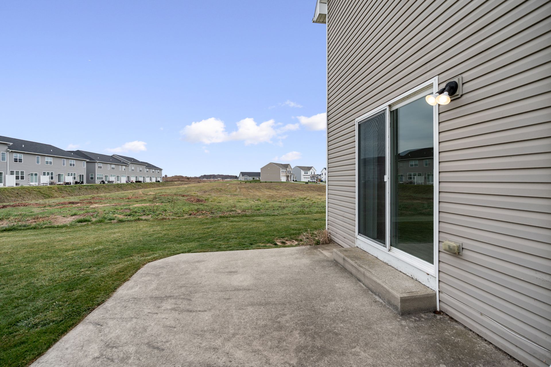 Backyard patio with sliding glass door and siding, facing open field and blue sky.