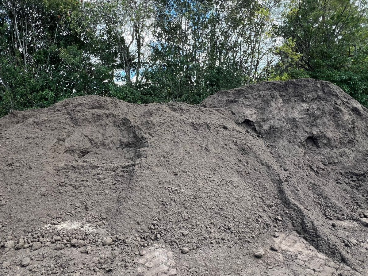 A large, tall pile of dark brown topsoil or dirt set against a backdrop of green trees — Laurieton Landscape Supplies In Laurieton, NSW