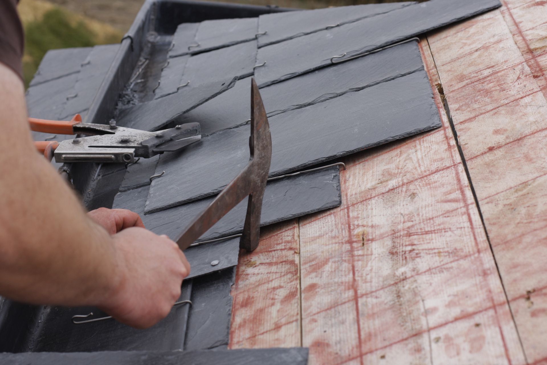 Worker installing dark roof shingles with a nail gun on a sloped roof, exposed underlayment visible
