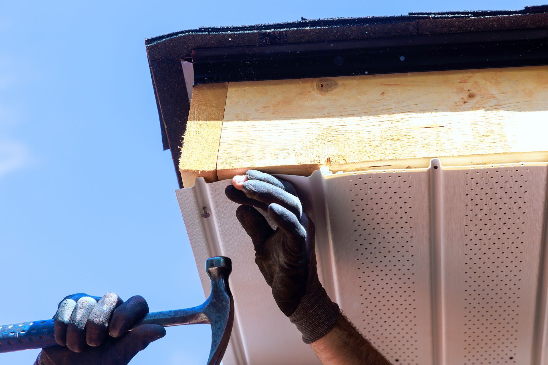 Gloved hands install a gutter on a house roof against a blue sky.
