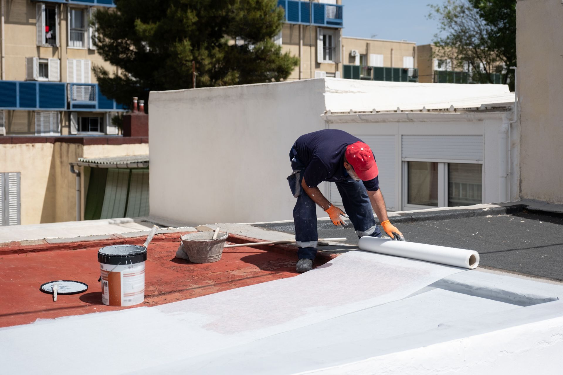 Worker installing a white roof membrane on a flat rooftop beside red roofing material.