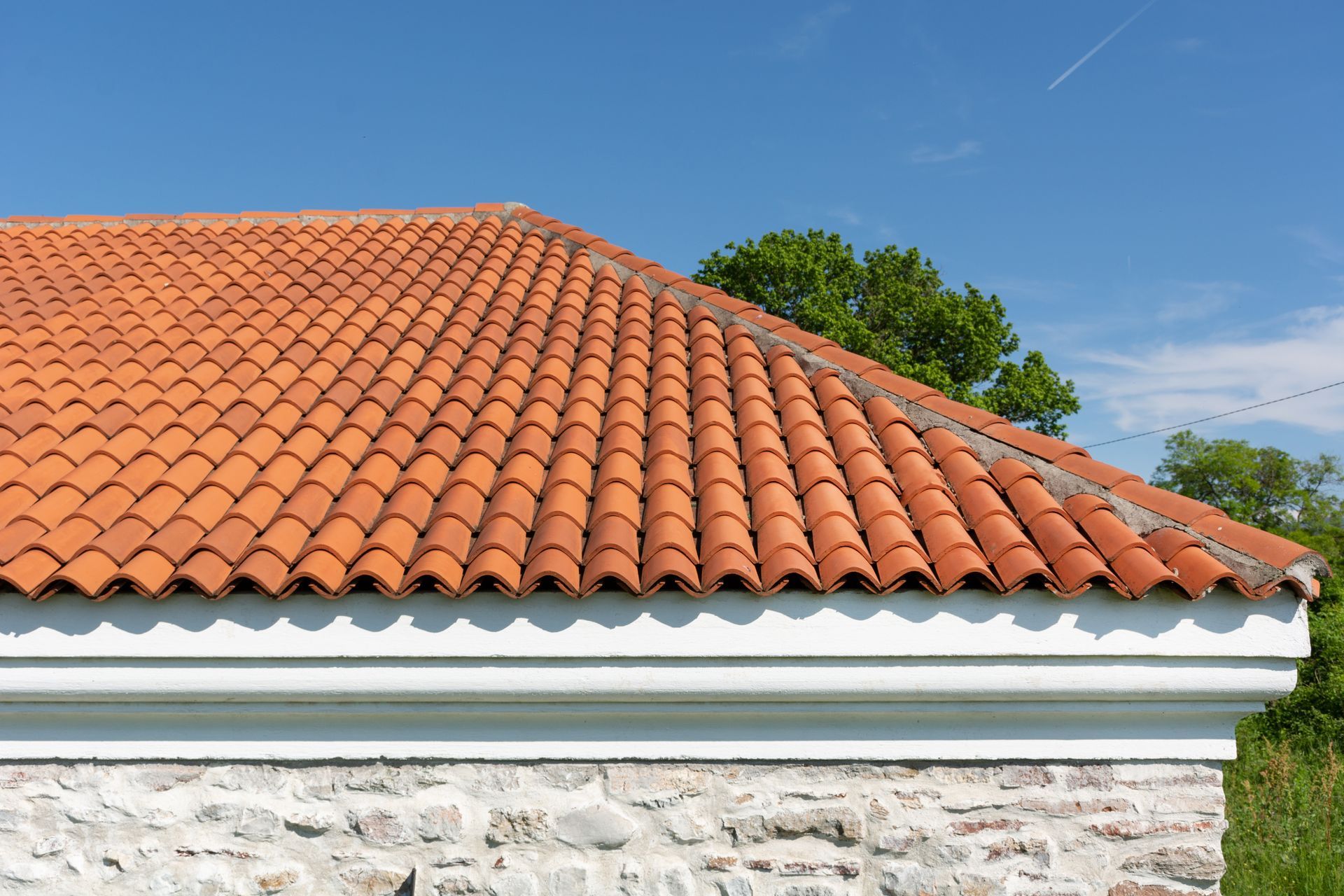 Terracotta-tiled roof above a white and stone wall, with trees and blue sky in the background