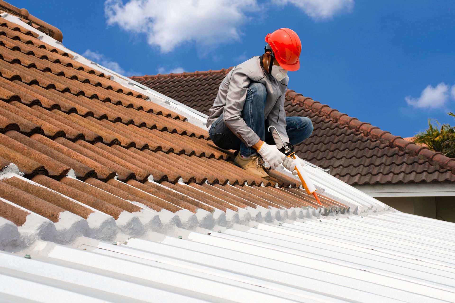 Worker in an orange hard hat spray-painting a white roof beside brown tiled roofing