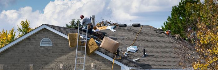 Roofers working on a damaged shingle roof with ladders, tools, and materials under a partly cloudy sky