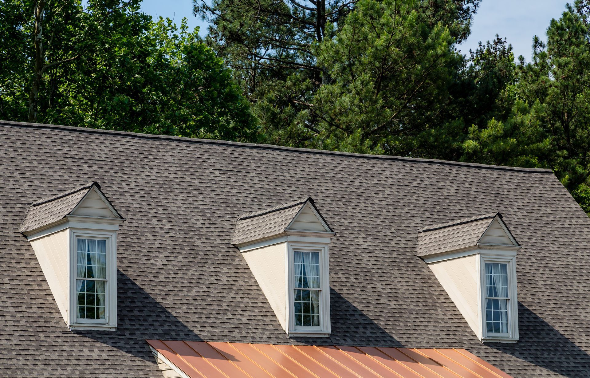 Three dormer windows on a gray shingled roof with trees in the background