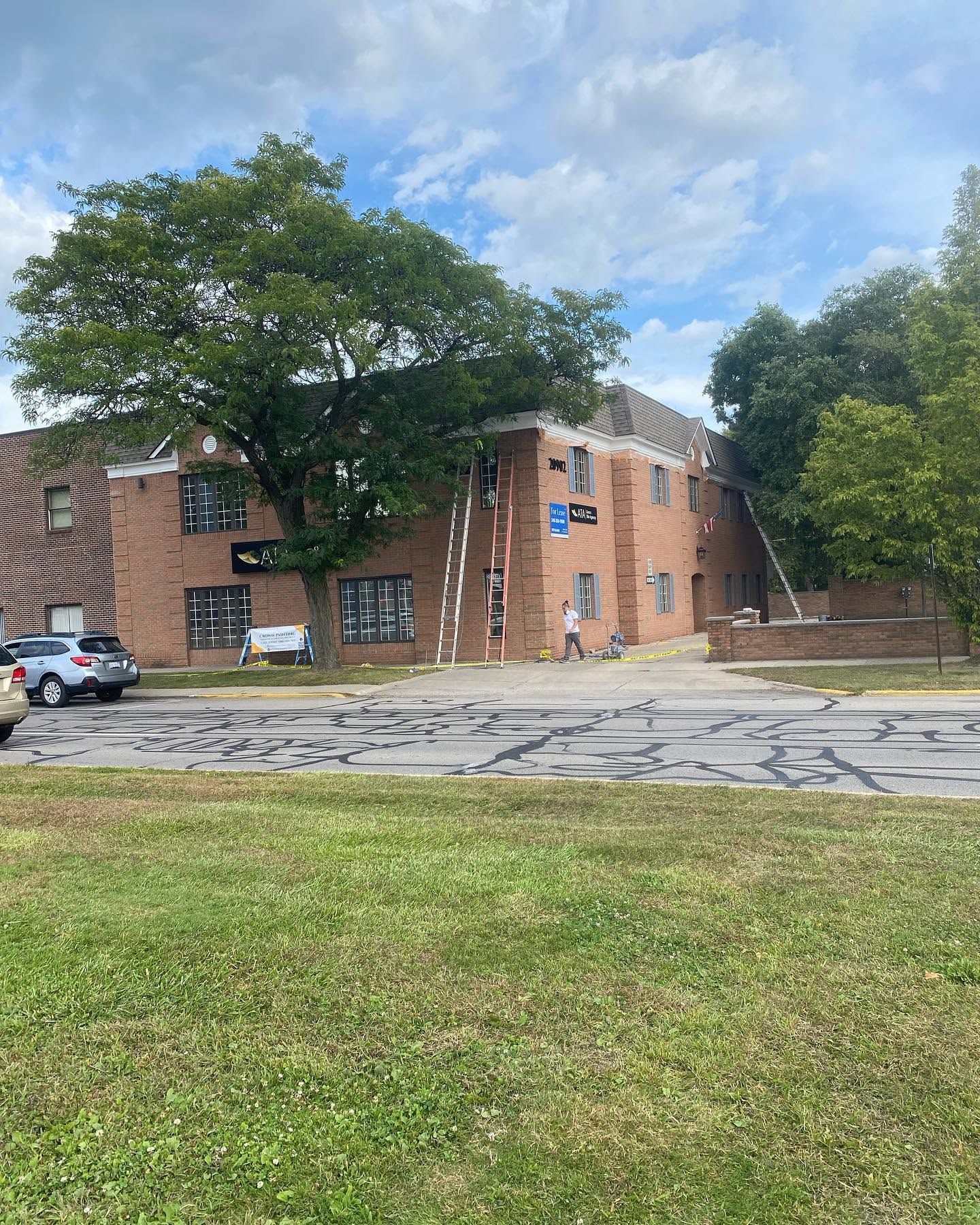 Brick apartment building with blue sign, parked cars, and large trees beside a cracked road and grassy foreground