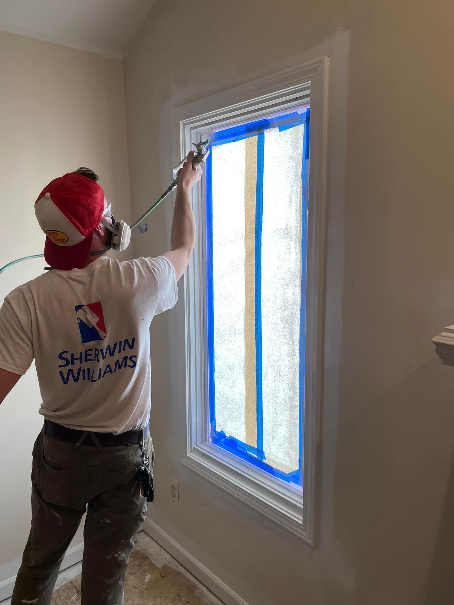 Painter taping off a window frame with blue tape in a room with beige walls.