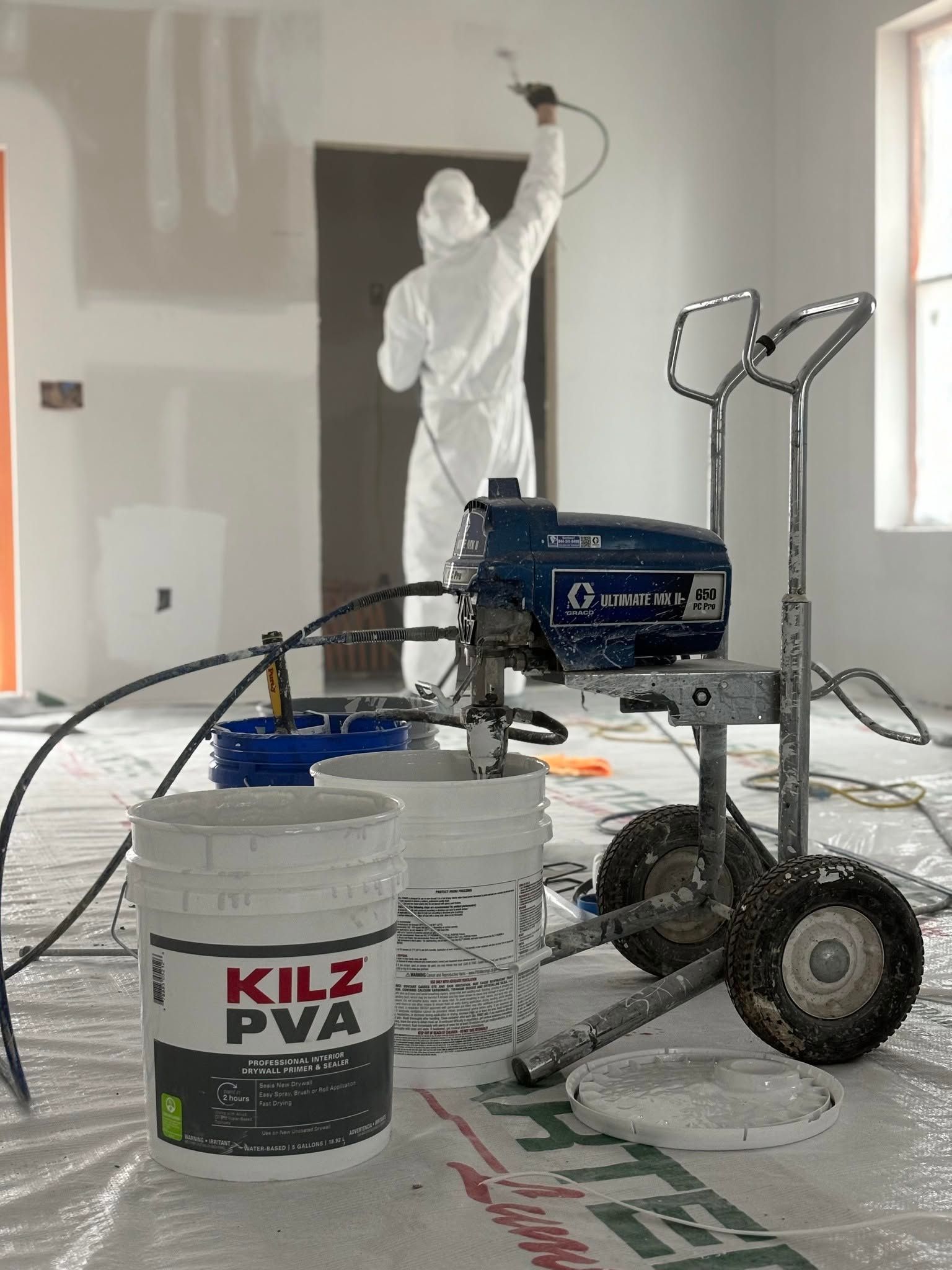 Painter in white coveralls spraying a wall with a paint rig beside buckets and a wheeled paint machine.