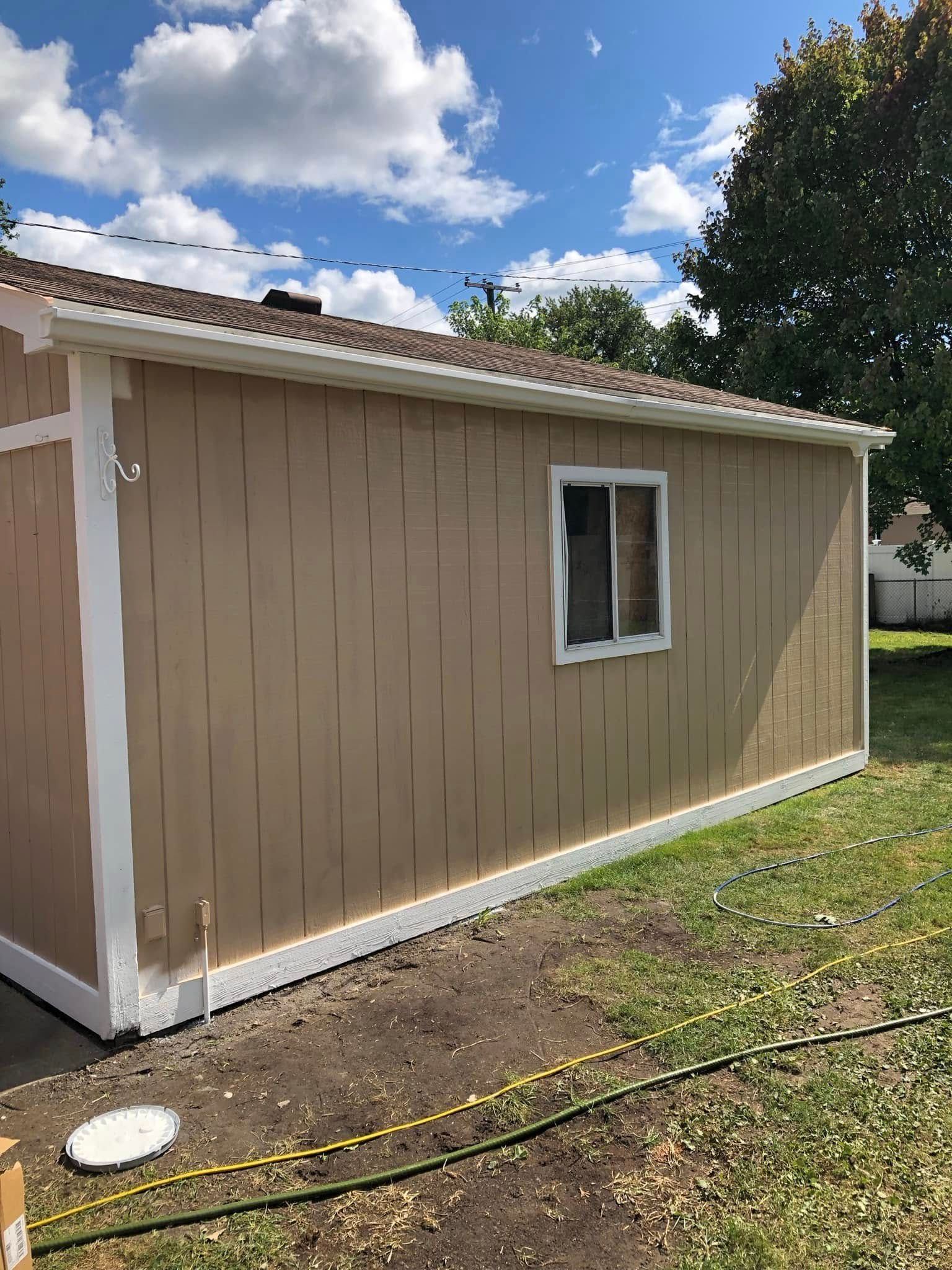 Beige shed with white trim and a small window in a grassy yard under a blue sky with clouds