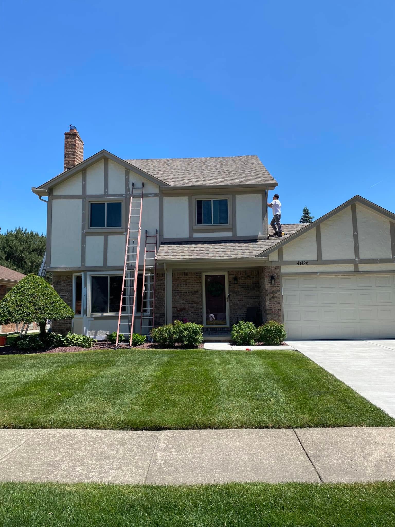 Two-story suburban house with beige siding, white garage, front lawn, and a ladder against the facade under a blue sky