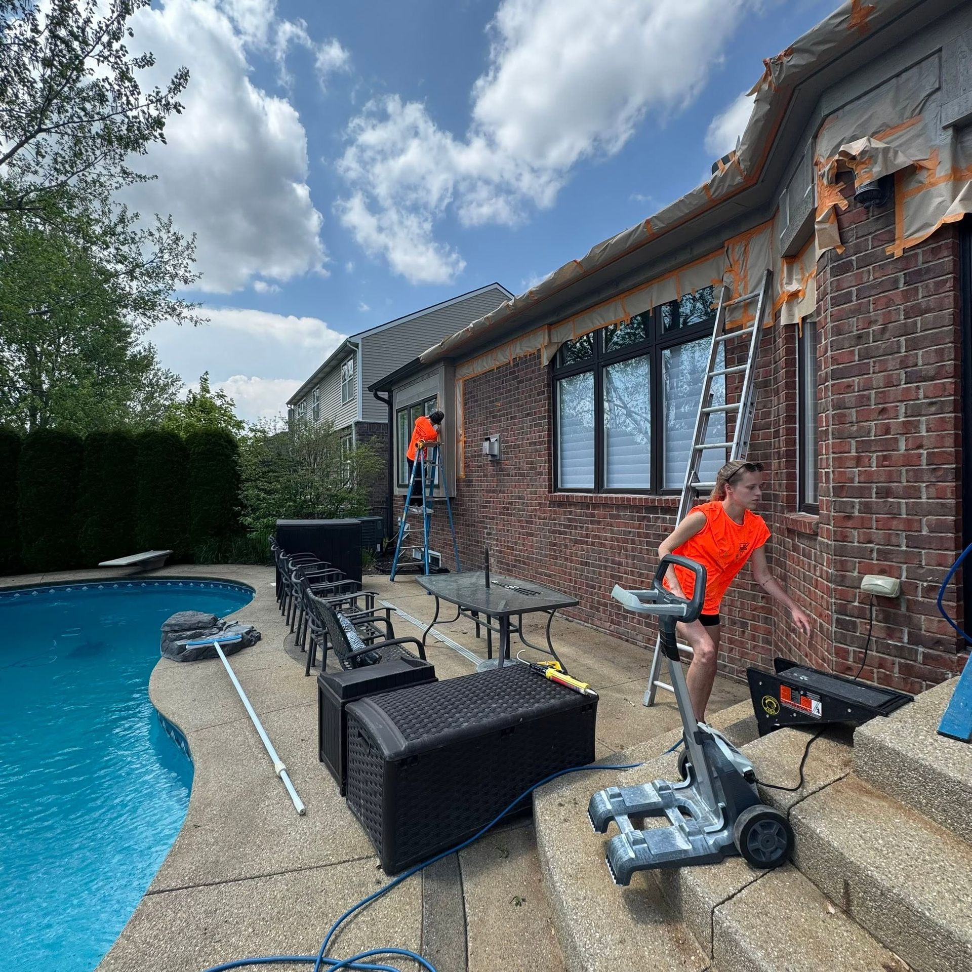 Workers on a brick house patio cleaning windows beside a pool under a cloudy sky.