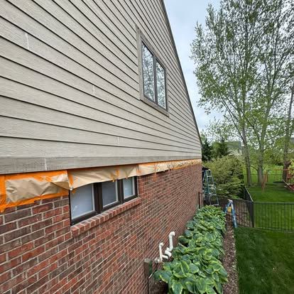 Brick-and-siding house exterior with exposed insulation under repair beside a green yard and trees