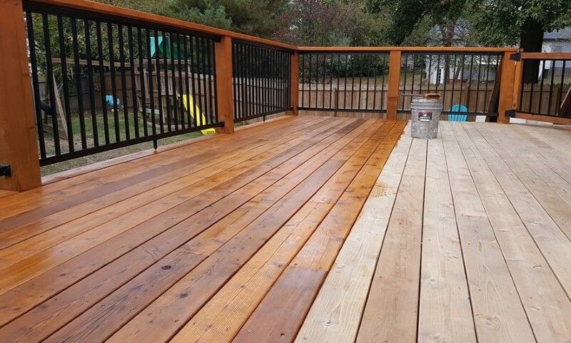 Wooden deck with black railings and one gray board section, overlooking a wooded area.