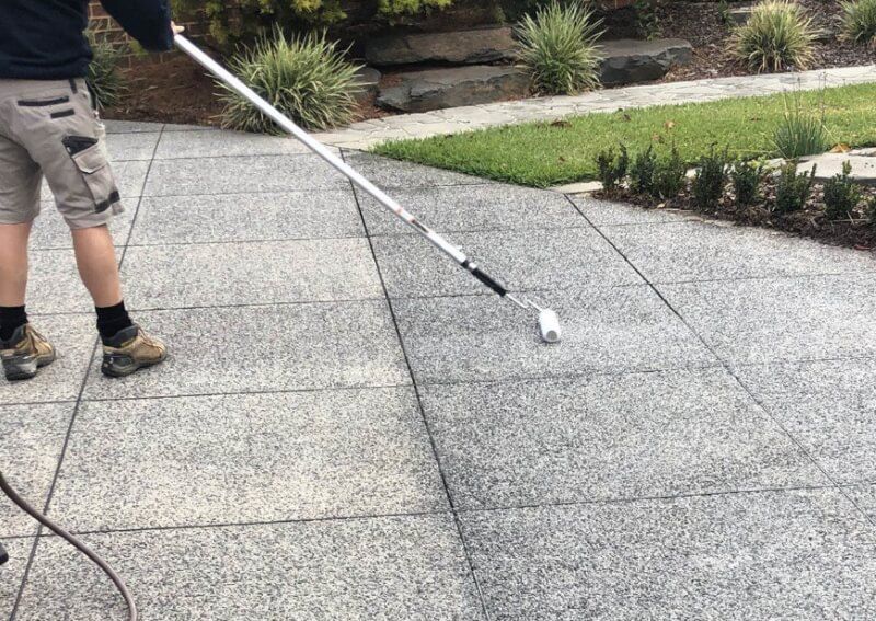 Person using a long-handled roller to coat a gray stone patio outdoors
