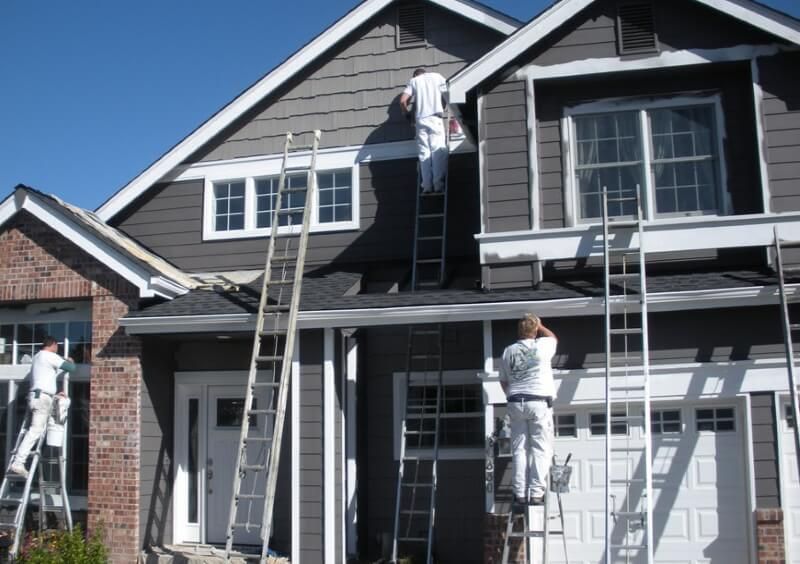 Workers painting the exterior of a gray two-story house on ladders