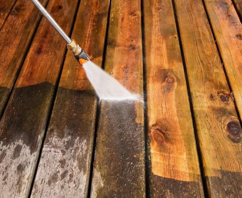 Pressure washer spraying water on a wooden deck, cleaning dark planks beside bright ones
