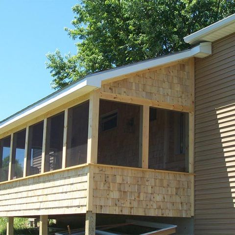 Wooden screened porch attached to a house with tan siding, viewed from the side under a blue sky.