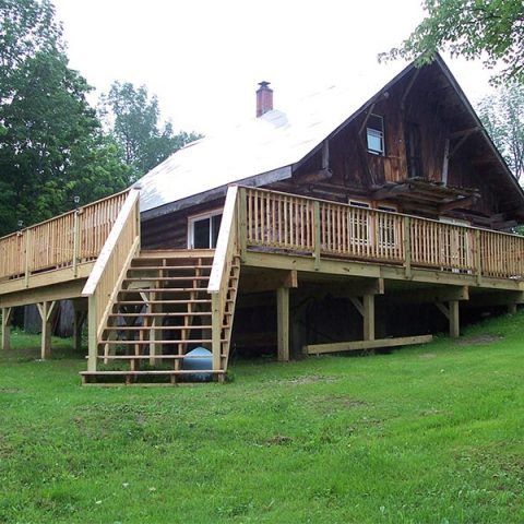 Wooden cabin on stilts with a wraparound deck and stairs, set on a grassy hillside.