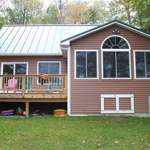 Suburban house with metal roof, front deck, and large windows, surrounded by a grassy yard and trees.