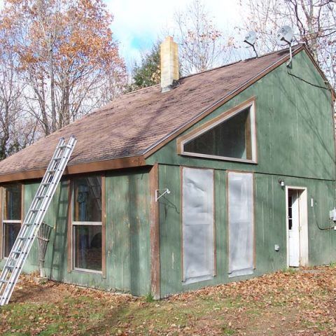 Green house with steep roof, ladder leaning outside, and autumn trees in the yard