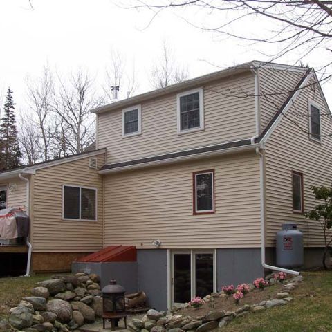 Two-story beige house with basement windows, stone landscaping, and bare trees in the yard