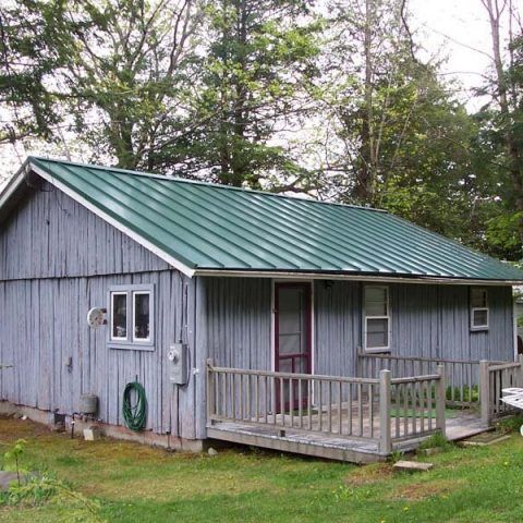 Small gray cabin with green metal roof and front porch, set in a wooded yard.
