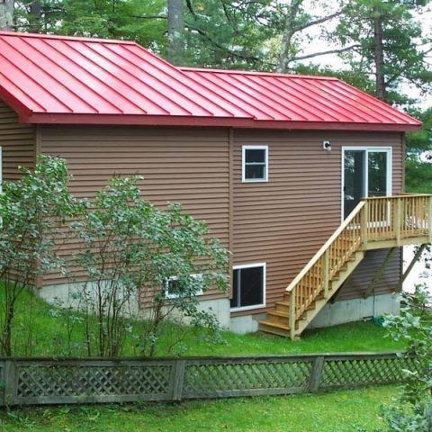 Brown house with a red metal roof, wooden stairs, and a small fenced yard amid trees