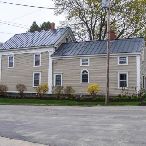 Two-story beige house with gray metal roofs and brick chimneys beside a paved road