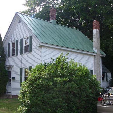 White house with green metal roof and brick chimneys, partially hidden by a large bush and trees.