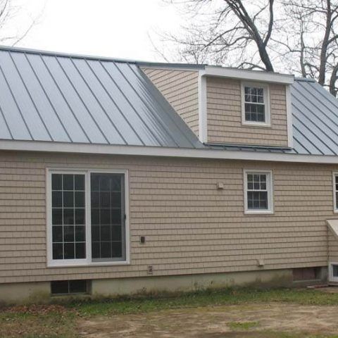 Beige house with metal roof, dormer window, and sliding glass doors under bare trees
