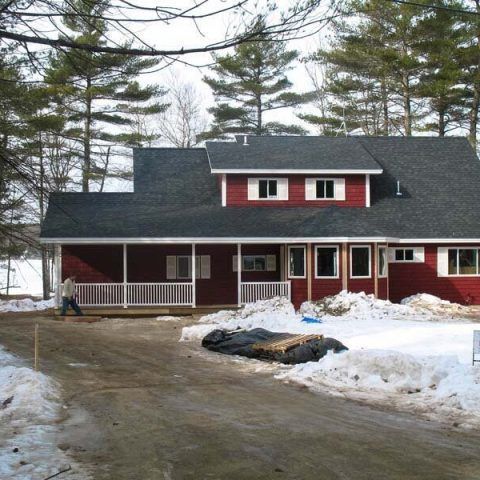 Red house with white trim in a snowy yard beside a driveway and tall trees