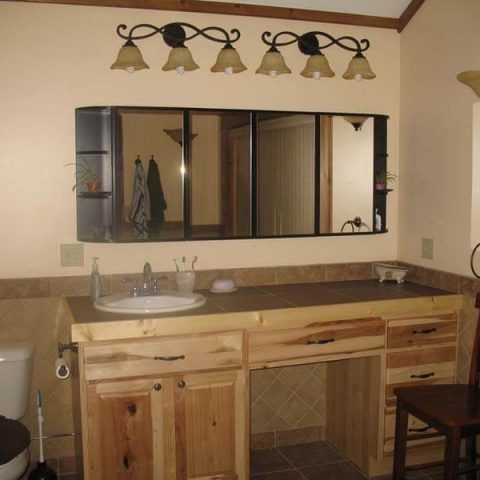 Wooden bathroom vanity with sink, mirror cabinet, and four brass light fixtures above beige tile countertop.