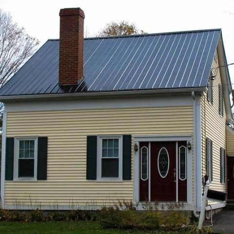Small beige house with dark metal roof, brick chimney, and red front door with white trim.