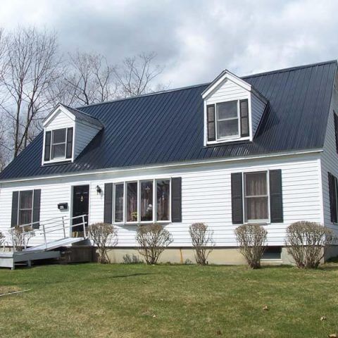 White house with dark blue roof, dormer windows, and a front ramp on a grassy lawn.