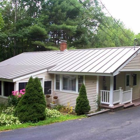 Single-story house with a silver metal roof, white siding, and a small porch beside a driveway and pine trees