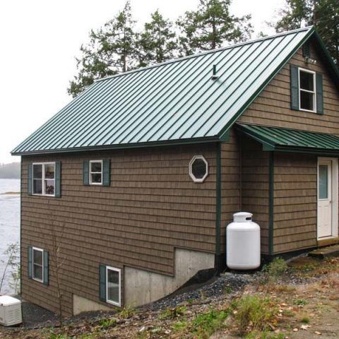 Small brown house with green metal roof and white propane tank by a rocky shoreline.