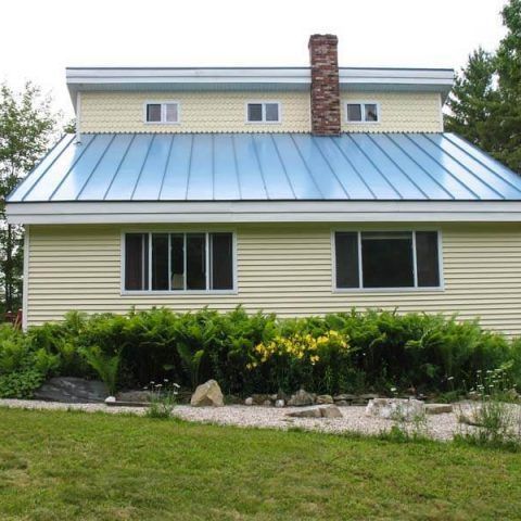 Single-story house with blue metal roof, brick chimney, and shrubs in front yard