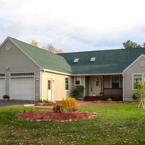 Single-story beige house with green roof, garage, and front porch on a grassy lawn