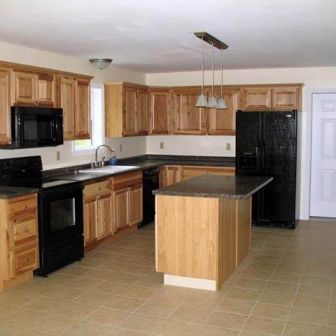 Empty kitchen with light wood cabinets, black appliances, center island, and tan tile floor.